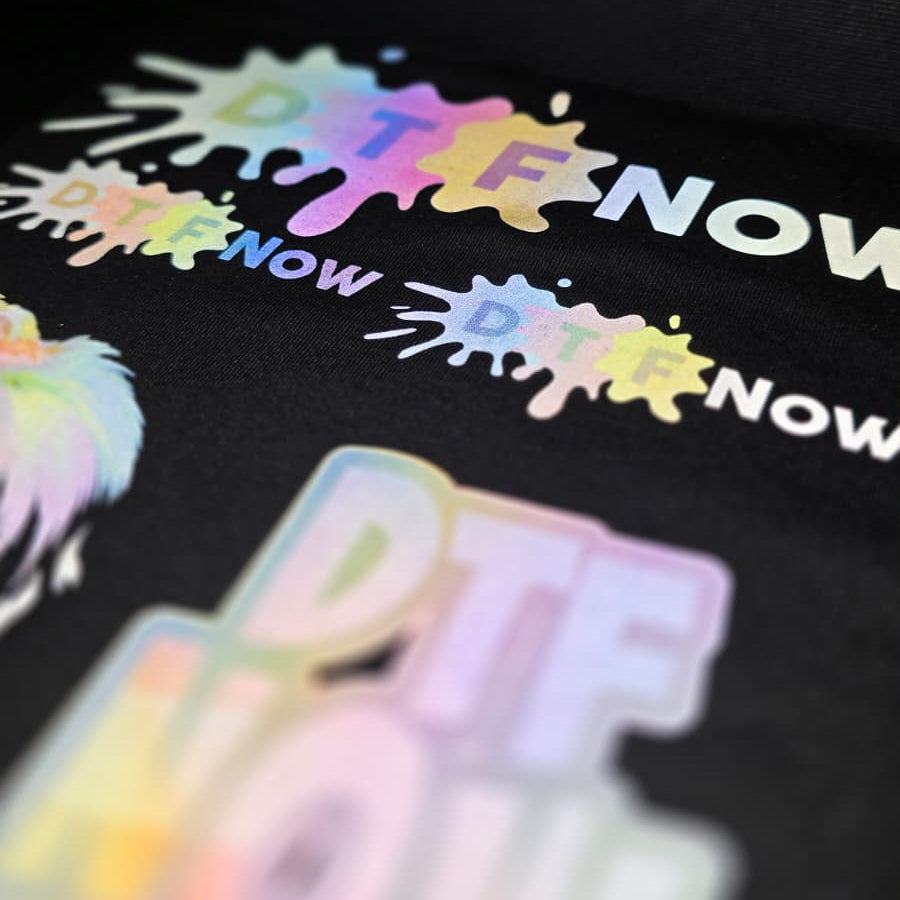 A close-up of a black surface featuring a colorful reflective spray-paint style 'DTF NOW!' graphic logo, with a blurred matching logo in the foreground.